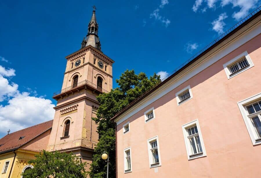 Kostol s vysokou vežou a historickým dizajnom, obklopený farebnými budovami a zelenými stromami, zac.