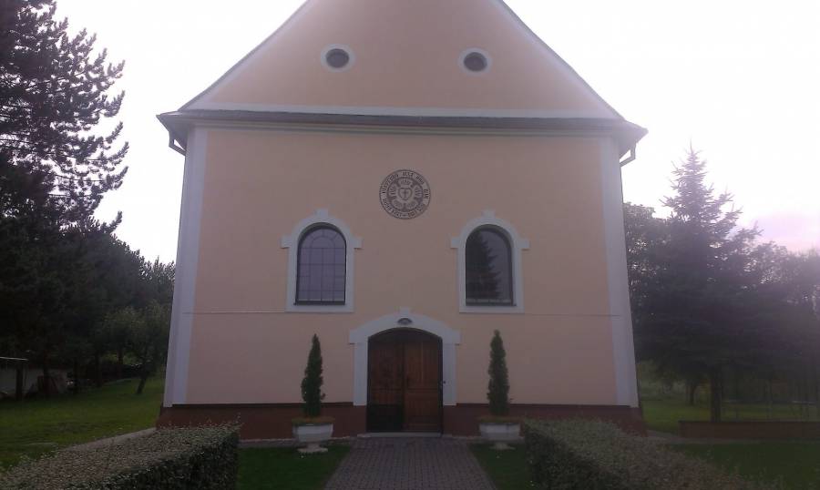 Kostol s jednoduchou architektúrou v prírodnom prostredí, obklopený stromami a záhradou.